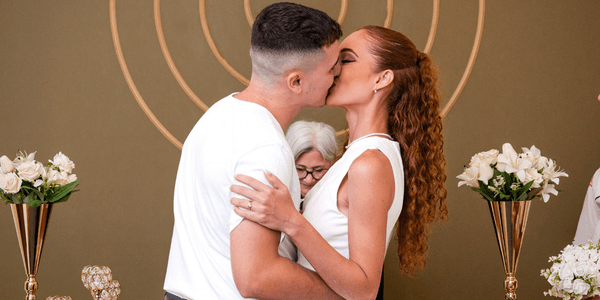 Casal se beijando e abraçado, com uma senhora ao fundo